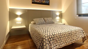 A bedroom with a patterned bedspread, a headboard with integrated lighting, a wall-mounted artwork, and a window with a view. - APARTMENT LA BOLERA INDOOR BOWLING ALLEY (Llanes)