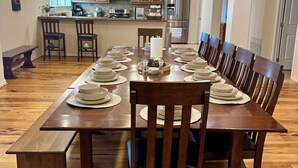 A dining area with a long wooden table set for a meal, flanked by wooden chairs. A bench is positioned at one end of the table. The room has wooden flooring and a kitchen area in the background with wooden cabinets and stainless steel appliances. - Luxury 90-acre 5BR Ranch near Cedar Creek (Kemp)