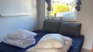 Ein Einzelbett mit blauer Matratze und weißen Kissen, ein Fenster mit Vorhängen und eine kleine Statue auf der Fensterbank.