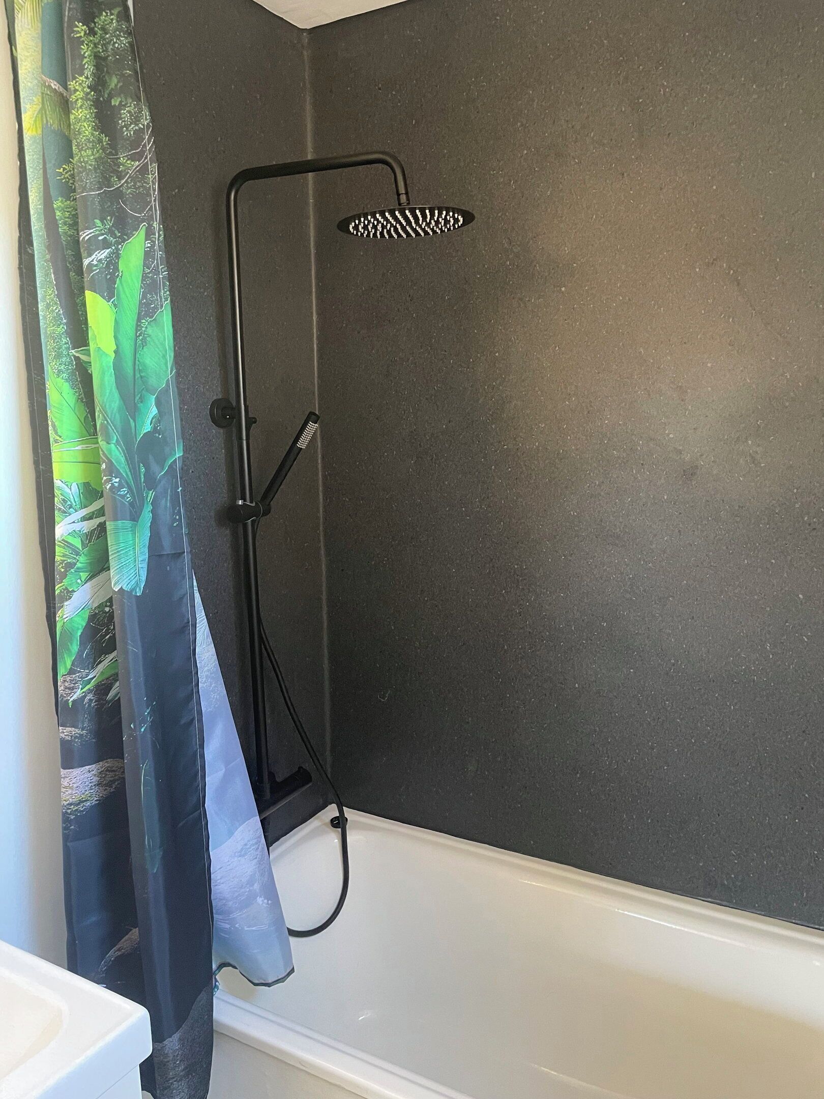Une douche avec un tuyau noir et un pommeau de douche, une baignoire blanche et un rideau orné d’un motif de feuilles vertes.