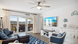 Ein geräumiges Wohnzimmer mit Deckenventilator, einem großen Fenster mit Vorhängen, einer Couch, einem Sessel und einem Fernseher an der Wand.