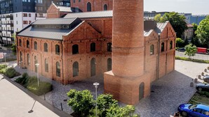 Un edificio histórico de ladrillo con una torre destacada, rodeado de edificios modernos y un área pavimentada con árboles y un automóvil.