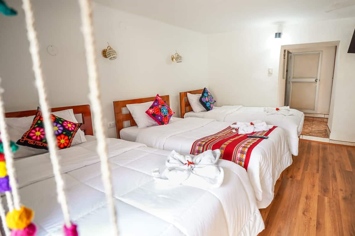 A hotel room with three beds, each with white bedding and colorful pillows. There are wall-mounted light fixtures and a wooden door.