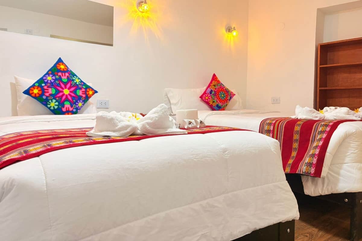 A hotel room with two beds, each with white linens and colorful pillows. There are towels on the beds and a small table with a cup and a book.