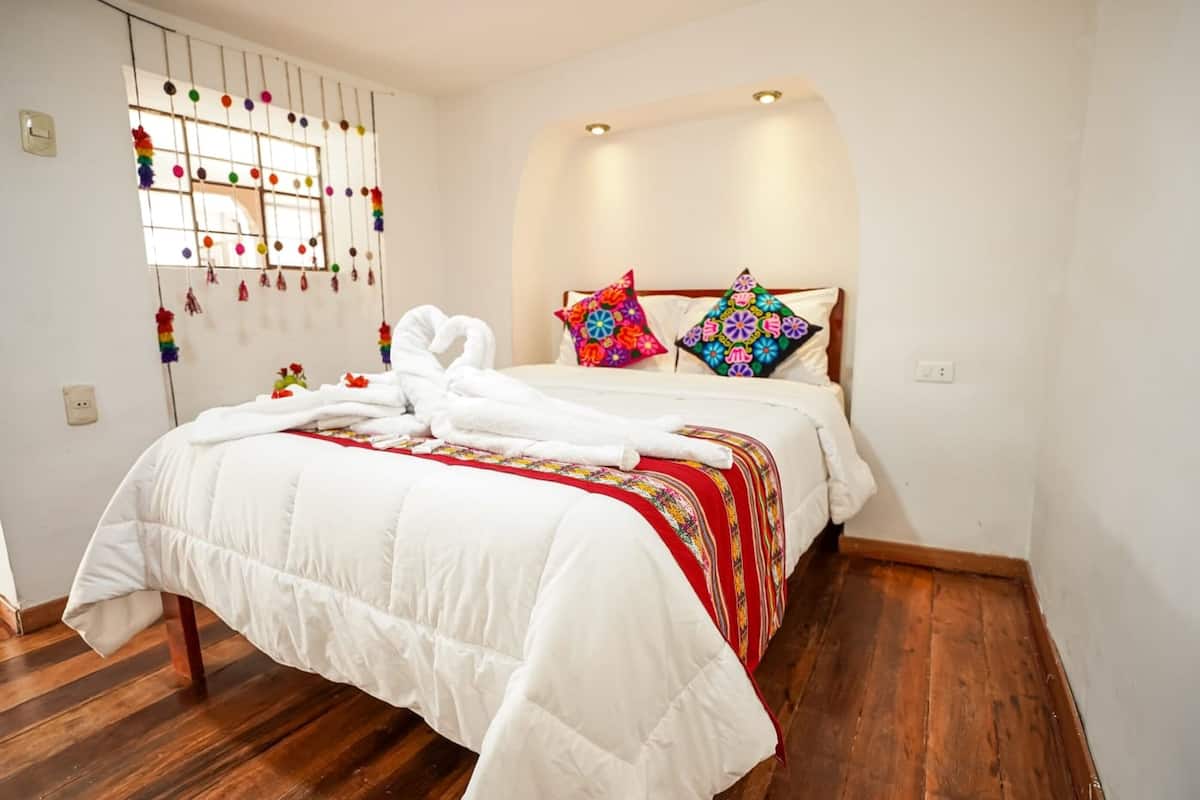 A neatly made bed with white bedding and colorful pillows, a wooden floor, and a window with a decorative curtain.