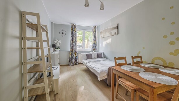 Een kamer met een bed, een houten trap, een bureau, een stoel, een tafel met placemats, een raam met gordijnen en een wandklok.