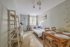 A room with a bed, a wooden ladder, a desk, a chair, a table with placemats, a window with curtains, and a wall clock.