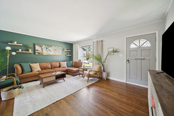 A living room with a sectional sofa, wooden floor, and a large window with curtains. - Earth & Echo-Nature-Inspired - Themed Bedrooms + Firepit + Walk to Brookside (Tulsa)
