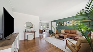 Un salon moderne doté d’un téléviseur à écran plat, de planchers en bois, d’un mur d’accent vert et d’un coin salon confortable avec un canapé et des fauteuils.