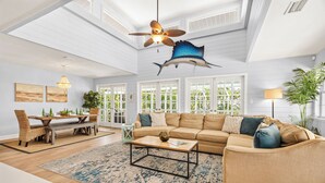 A spacious living room with a high ceiling, a ceiling fan, a large fish decoration, a sofa set, a coffee table, and a dining area with wicker chairs. - Salty Sailfish | Private Pool + Hot Tub + Siesta Key (Sarasota)