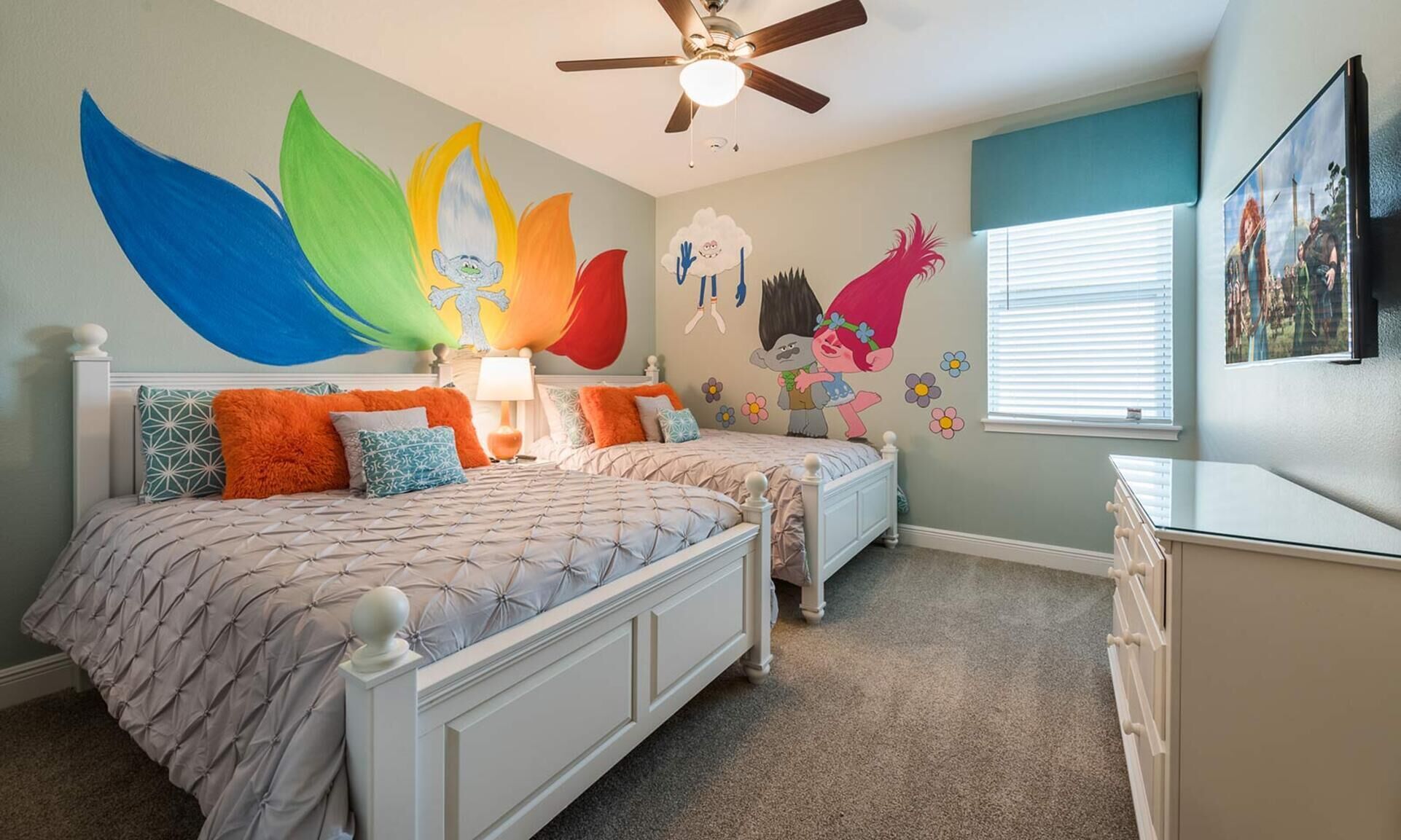 A bedroom with two beds, a ceiling fan, a window with blinds, and a wall with colorful wall art.
