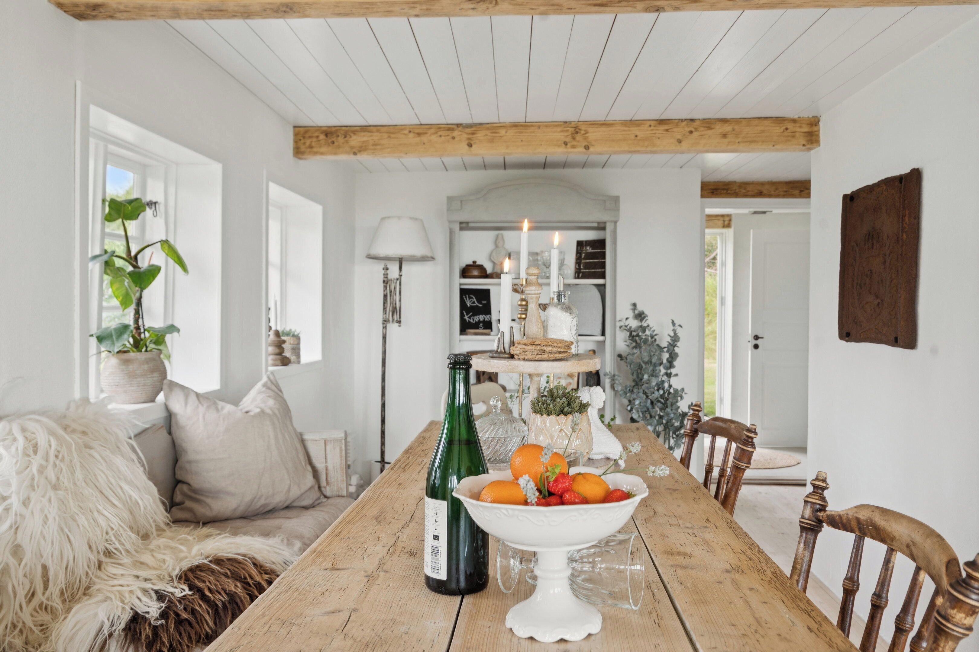 A wooden dining table with a bowl of fruit and a bottle of wine, a white sofa with a fur throw, and a plant in a pot.