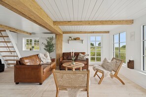 Ein geräumiges Loft mit hohen Decken, Holzbalken, einer Treppe, einem braunen Ledersofa, einem Korbstuhl und Blick ins Grüne durch große Fenster.