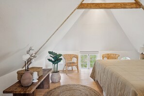 Une chambre mansardĂ©e chaleureuse, avec un plafond en pente, un lit, deux fauteuils en osier, une petite table sur laquelle sont posĂ©s un vase et une bougie, et une fenĂȘtre donnant sur un paysage verdoyant.