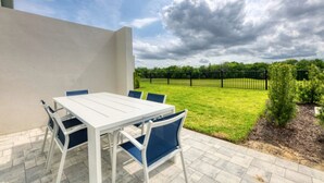 Een terras buiten met een witte tafel en blauwe stoelen, een gazon en een zwarte hek in de achtergrond.
