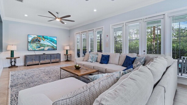 A spacious living room with a large sofa, a coffee table, and a ceiling fan. There are large windows with blinds, a TV mounted on the wall, and a cabinet with a decorative top. - Embrace Luxury: Your Perfect Escape, Orlando Villa 1000 (Kissimmee)