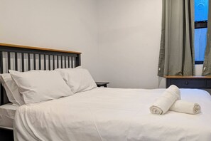 Una cama bien tendida con sábanas y almohadas blancas, cabecera de madera, una mesita de noche pequeña y una ventana con cortinas.