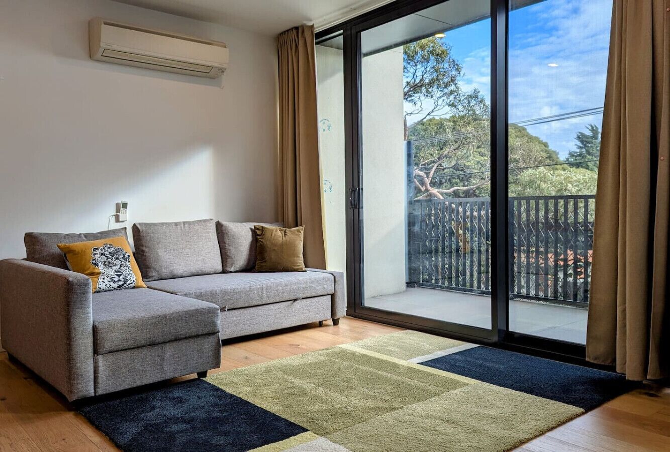 A modern living room with a grey sofa, a patterned throw pillow, and a large sliding glass door leading to an outdoor balcony.