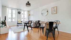 A modern living room with a sofa, dining table, chairs, and a coffee table. There are large windows with curtains, a ceiling light, and wall art.
