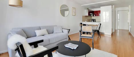 Een moderne woonkamer met een grijze bank, een zwarte salontafel en een eethoek, met een keuken op de achtergrond.