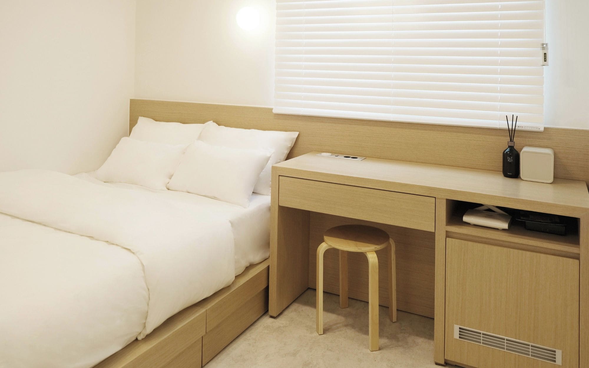 A bedroom with a bed, a desk, a stool, and a window with blinds.