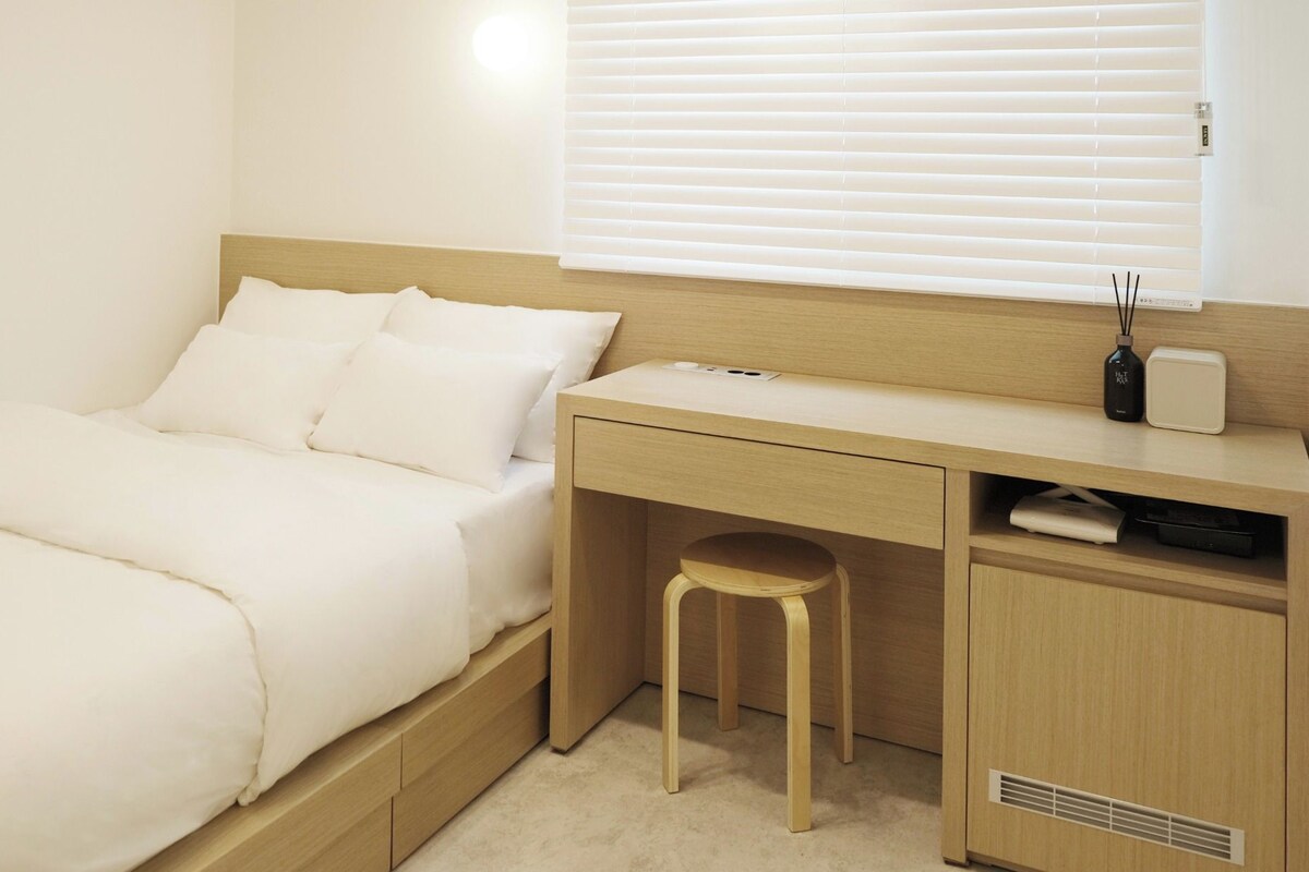 A bedroom with a bed, a desk, a stool, and a window with blinds.