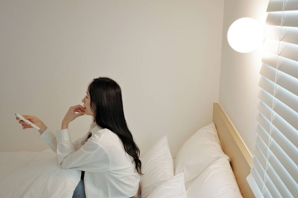 Una persona sentada en una cama con ropa blanca, usando un teléfono inteligente.