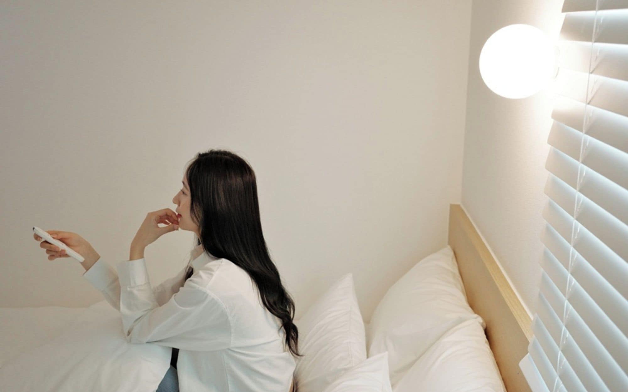 Eine Person sitzt auf einem Bett mit weißer Bettwäsche und hält eine Fernbedienung.
