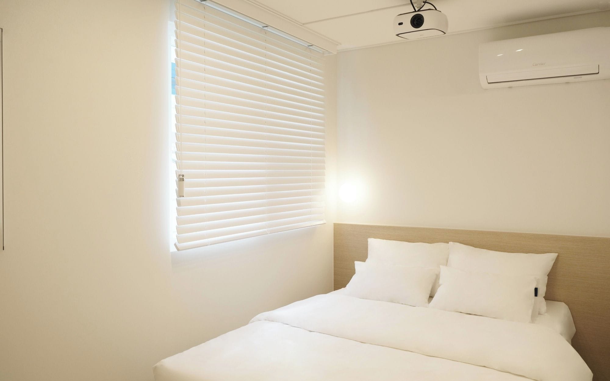 Une chambre d’hôtel avec un lit, du linge de lit blanc, une fenêtre avec des stores, un appareil de climatisation et un projecteur fixé au plafond.