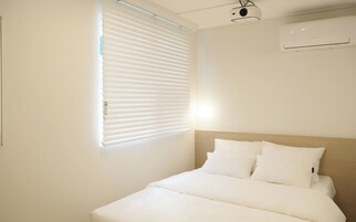 Une chambre d’hôtel avec un lit, du linge de lit blanc, une fenêtre avec des stores, un appareil de climatisation et un projecteur fixé au plafond.