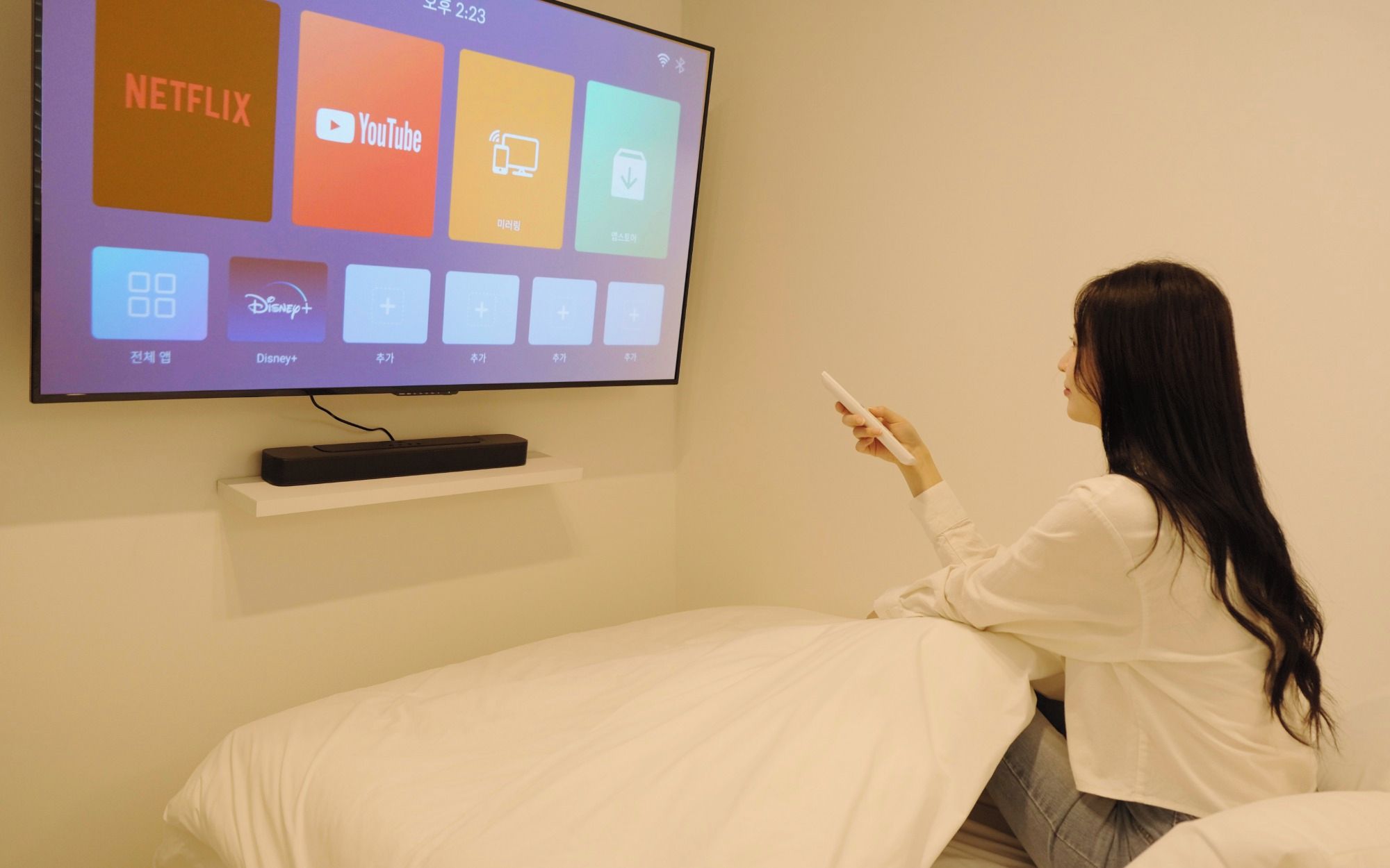 Una persona sentada en una cama en una habitación con un televisor montado en la pared que muestra diferentes servicios de streaming.
