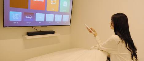 Una persona sentada en una cama en una habitación con un televisor montado en la pared que muestra diferentes servicios de streaming.