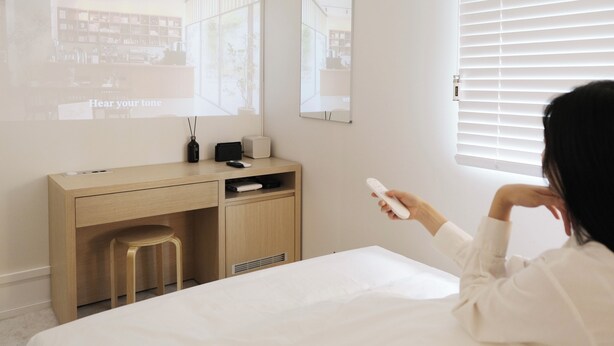 A bedroom with a bed, a desk, and a chair. There is a window with blinds, a mirror on the wall, and a projector screen displaying "Hear your tone". - Bluenote (Incheon)