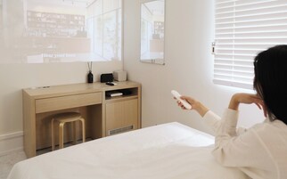 A hotel room with a bed, a desk with a chair, a window with blinds, and a person holding a remote control.