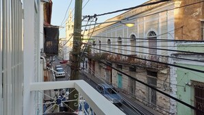 Un balcon avec un fauteuil en osier et un coussin rouge, donnant sur une rue animée par la circulation des voitures et bordée d’immeubles.