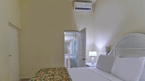 Une chambre à coucher avec un lit, une table de chevet avec une lampe, une porte blanche et une salle de bain visible à travers une porte ouverte.