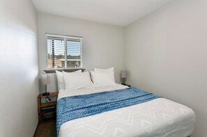 Una cama bien hecha con sábanas blancas y una manta azul, una mesita de noche con lámpara, y una ventana con persianas.