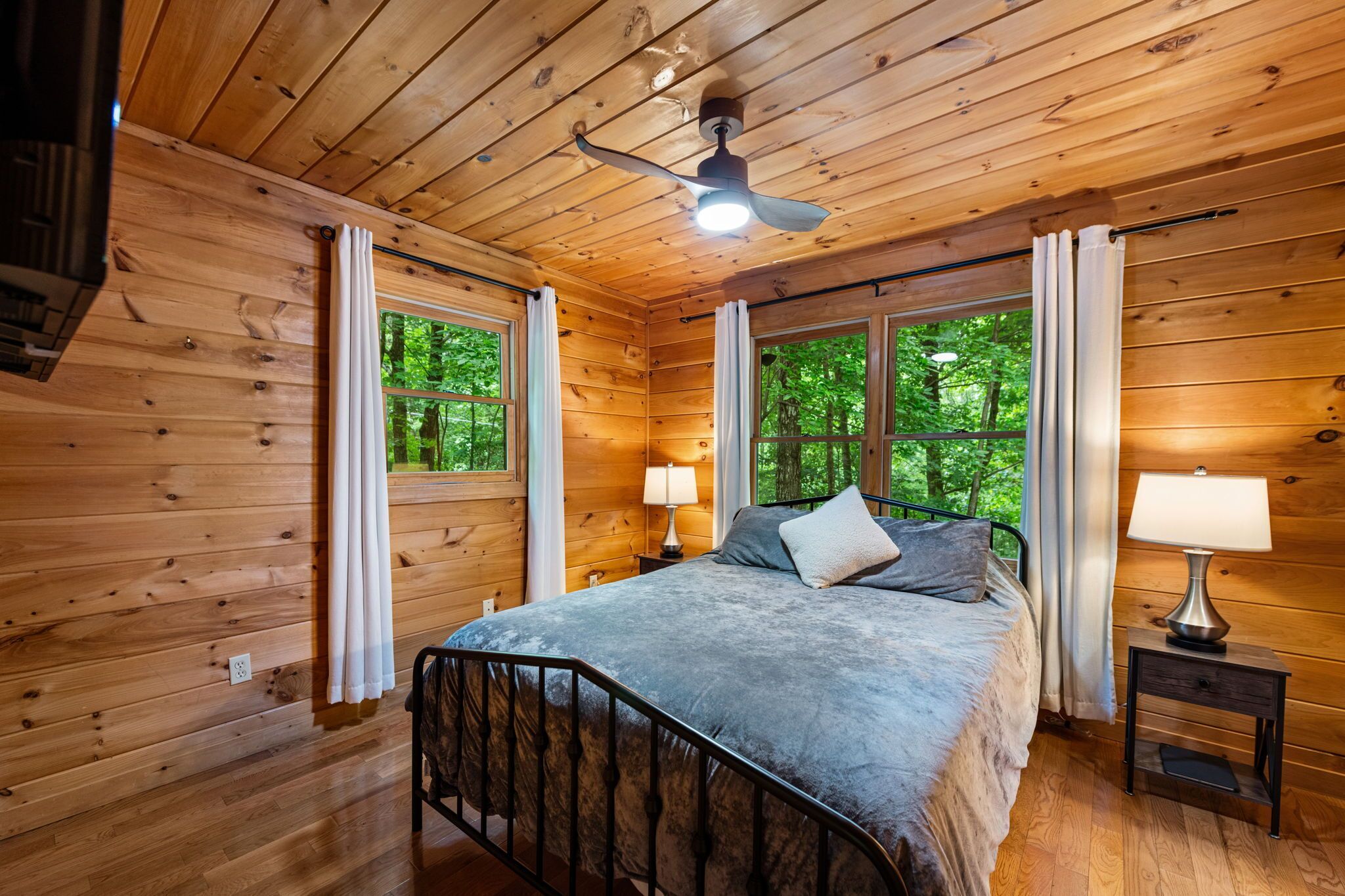 Une chambre à coucher dans une cabane en bois, avec un lit, deux tables de chevet avec des lampes, un ventilateur de plafond et de grandes fenêtres avec des rideaux.