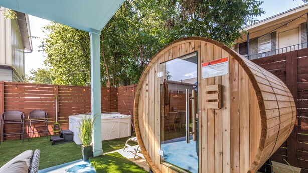 A wooden hot tub with a glass door, surrounded by a patio with artificial grass, outdoor furniture, and a wooden fence. - The Q - Austin Spa, Sauna, Gym, Backyard 7 Bed UT Home ATX (Austin)