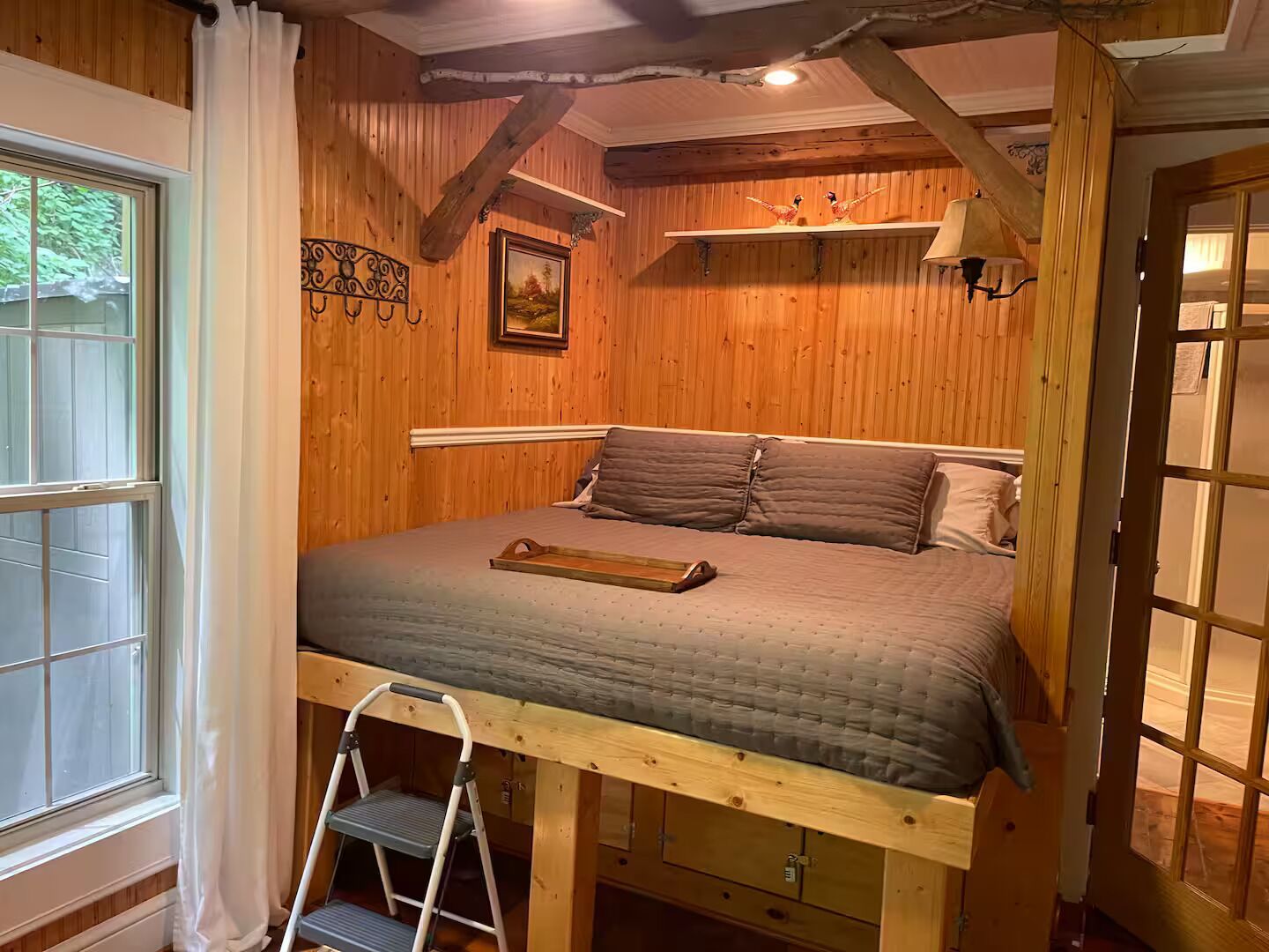 A wooden-paneled bedroom with a bed, a ladder, a painting, and a window.