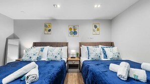 Une chambre d’hôtel avec deux lits, chacun agrémenté de draps et d’oreillers bleus assortis. Il y a deux tables de chevet avec des lampes et un miroir. Les murs sont blancs, et trois œuvres d’art encadrées sont accrochées au-dessus des lits.