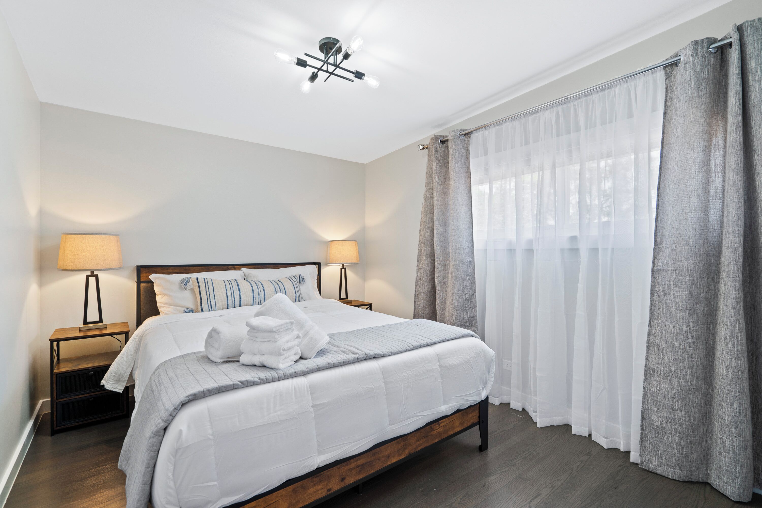 Una cama bien tendida con sábanas blancas y una manta gris, dos lámparas de noche, un cabecero de madera y cortinas grises traslúcidas.