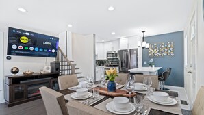 A modern dining room with a wooden dining table set for six, a flat-screen TV mounted on the wall, and a kitchen in the background.