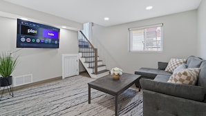 A modern living room with a flat-screen TV mounted on the wall, a grey sofa, a wooden coffee table, and a staircase.