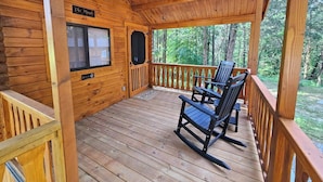 A wooden cabin with a covered porch, a ceiling fan, and a window overlooking a forest.