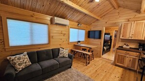 A cozy cabin interior with a wooden floor, a sofa with patterned cushions, a dining table, and a kitchen area with wooden cabinets.