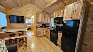A wooden cabin interior with a kitchen area featuring wooden cabinets, a black refrigerator, a microwave, and a dining table with stools.