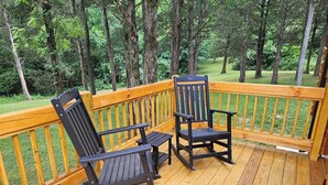 A wooden deck with two rocking chairs and a small table, surrounded by a wooden railing and a forested background.