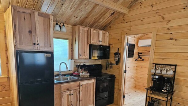 A compact kitchen with wooden cabinets, a black refrigerator, a microwave, and a coffee machine on a small shelf.