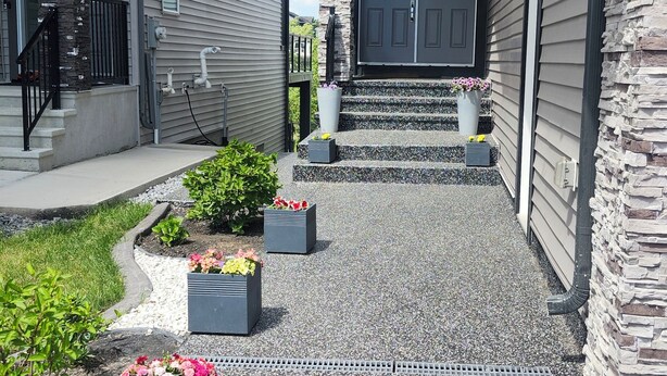 A residential driveway with a gravel surface, bordered by flower planters and a stone wall. - Charming 1-bedroom walkout pond backing suite with fitness machine, AC and bbq (Beaumont)